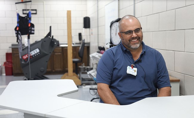 A man at a desk with exercise equipment behind him.