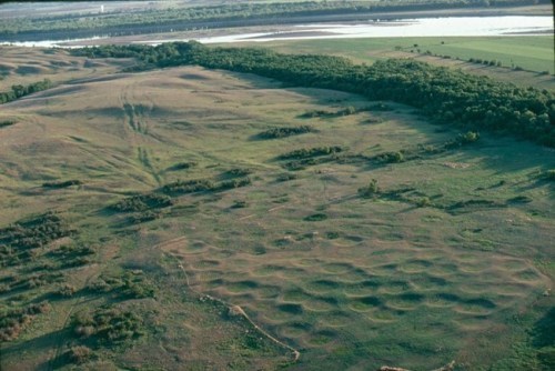 Aerial view of an open, grassy landscape near a river, patterned with shallow round depressions