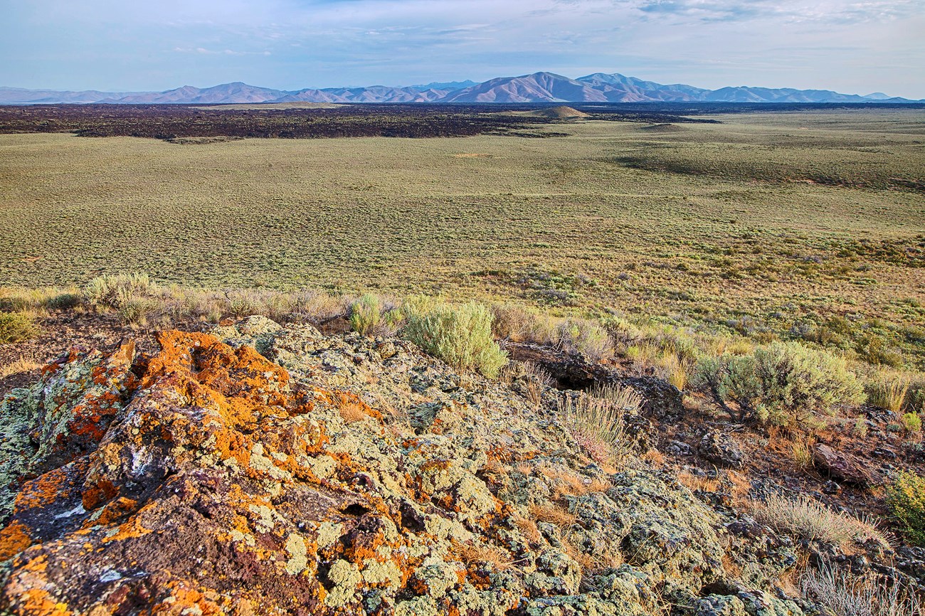 A vast, flat sagebrush landscape with dark lava rock and distant mountains.