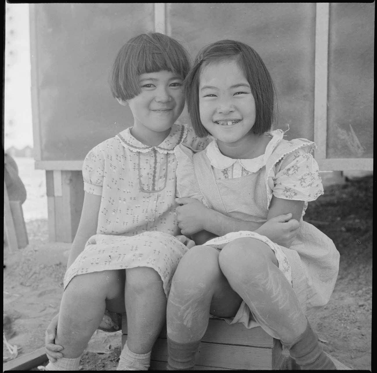 Two smiling young Japanese American girls in print dresses sitting close together on a wooden box