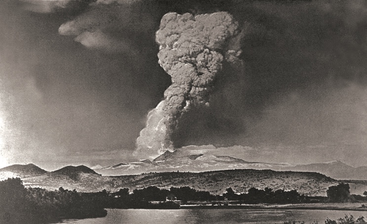 Black-and-white photograph of a huge mushroom-shaped eruption column rising above a distant mountain and a flat valley with a river in the foreground