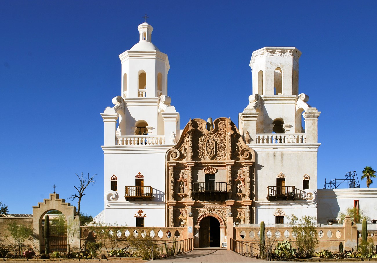 A long stone wall with an archway in front of a white mission church with two bell towers and an ornate brown facade.