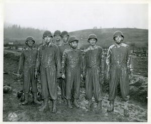 Seven helmeted soldiers covered in mud from the neck down, standing in a muddy field.