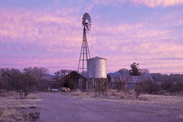 The colored clouds dot the sky as the sun rises over historic farming structures like a windmill, a water storage tank, and a barn in Las Cienegas National Conservation Area.