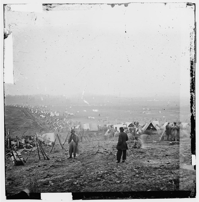Two men on a bare hilltop beside stacked rifles, with army tents in the distance.