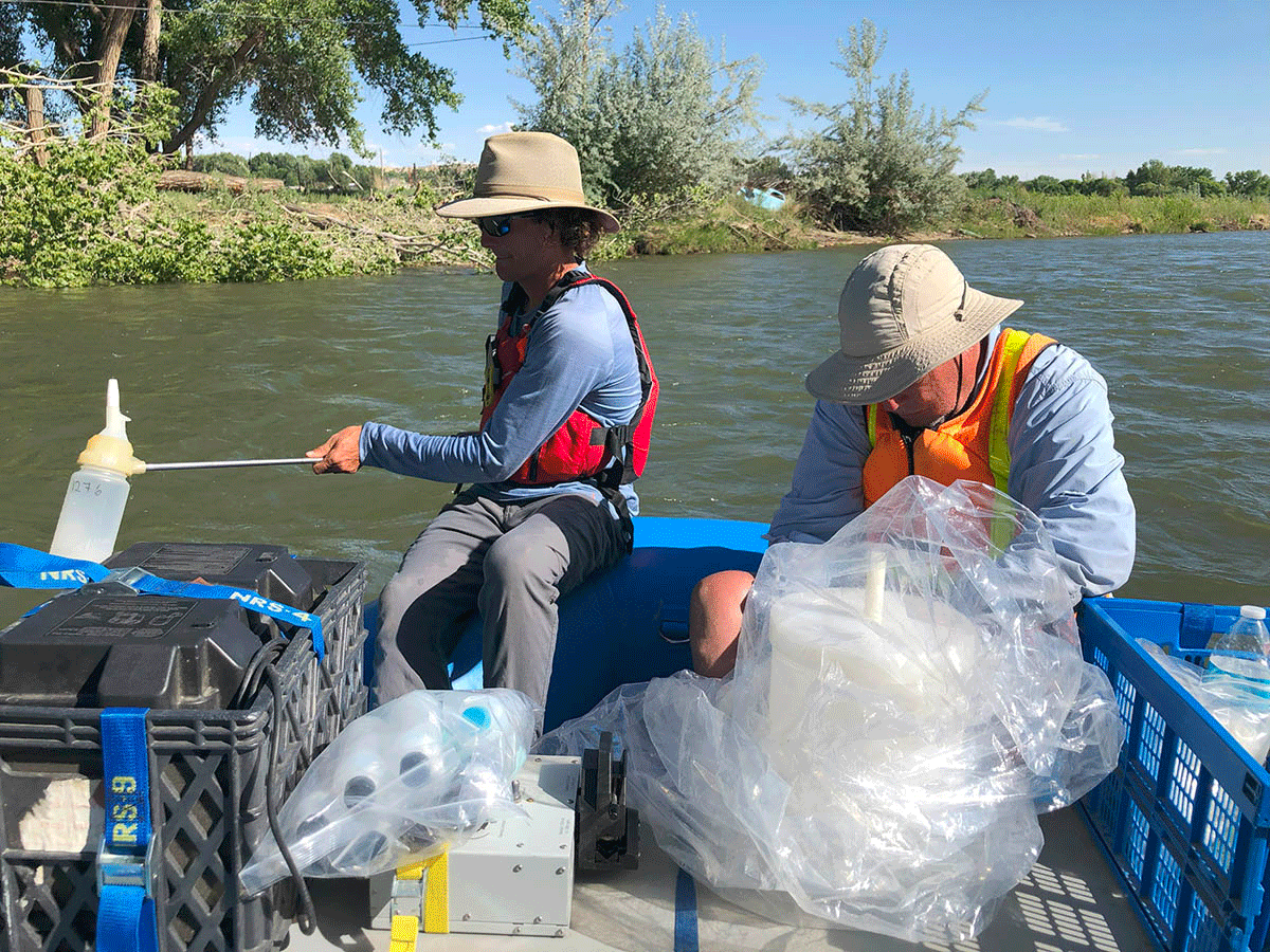 Two people in a boat on a river, one holding a water sampler.