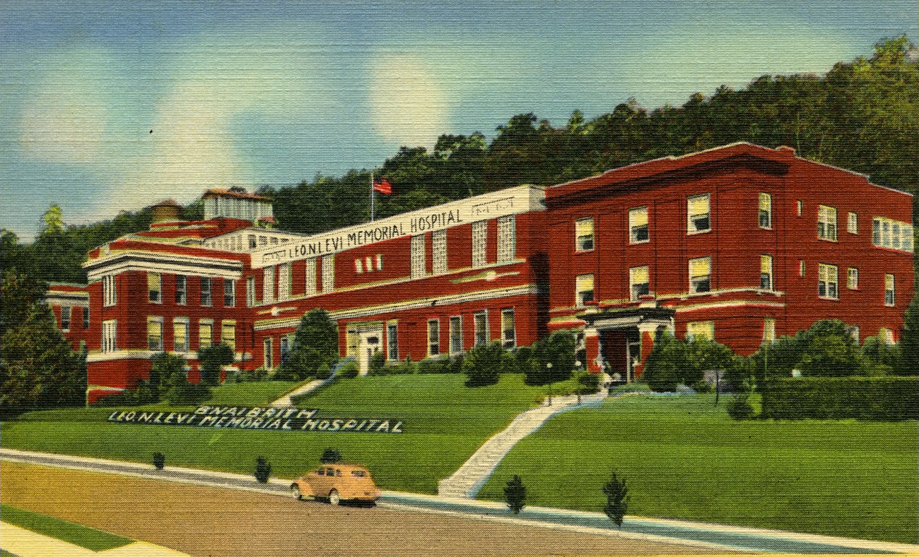 A colour postcard of a red-brick hospital on a hillside, with a yellow car on the road in front.