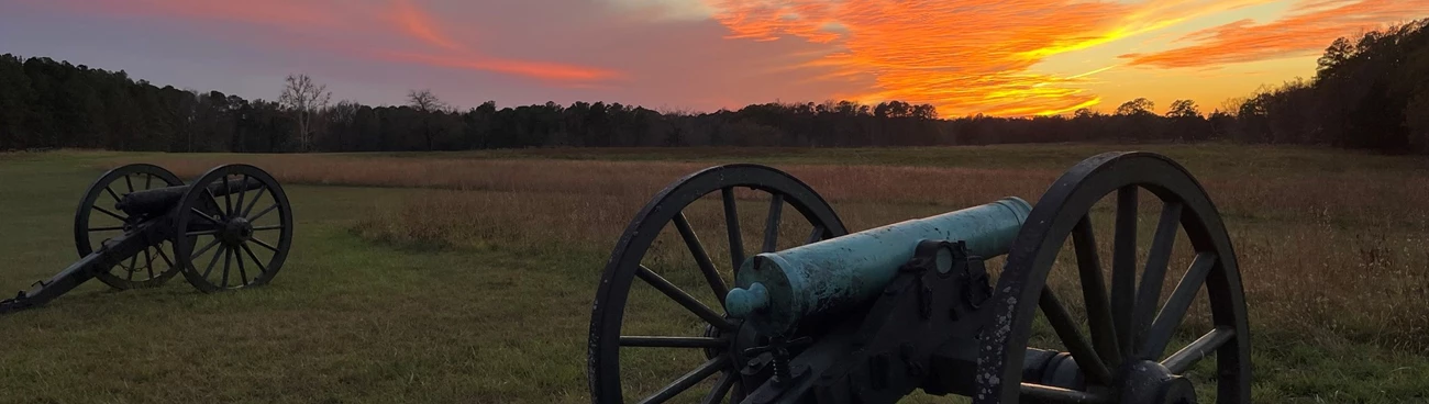 Civil War cannons in a grassy field under a vivid orange and pink sunset.