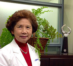Portrait of Dr. Alice Yu wearing a white physician's coat in her office, with plants visible through a window behind her.