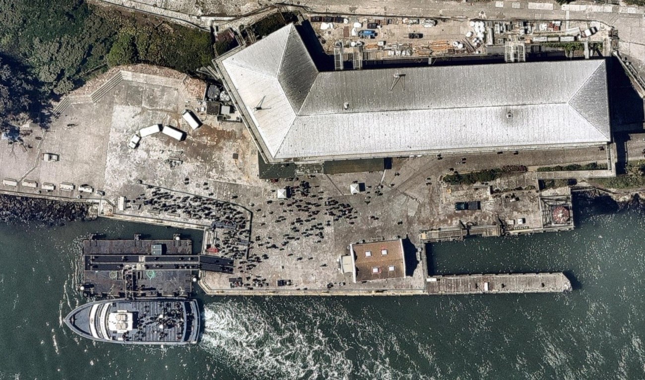 Aerial view of the Alcatraz concrete wharf, with a white ferry docked and a large grey-roofed building above