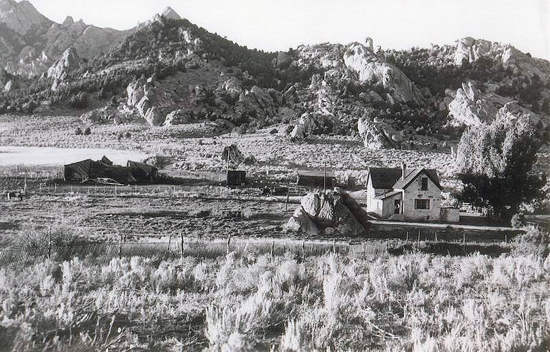 A rustic stone farmhouse in a field below rocky mountains.