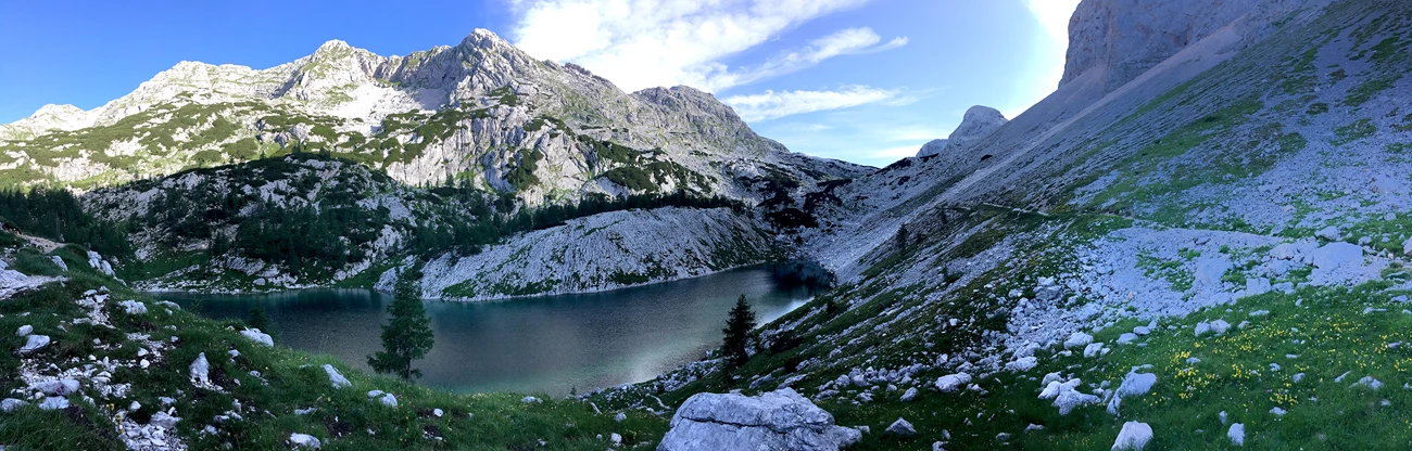A blue-green alpine lake among grey rocky mountains, green meadows and trees