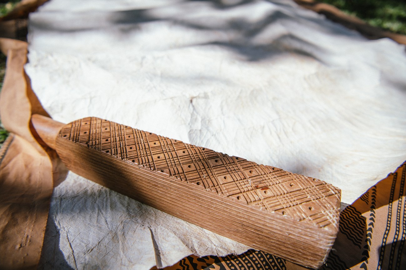 A rectangular wooden beater with diagonal carved lines resting on barkcloth that bears the imprint of its design.