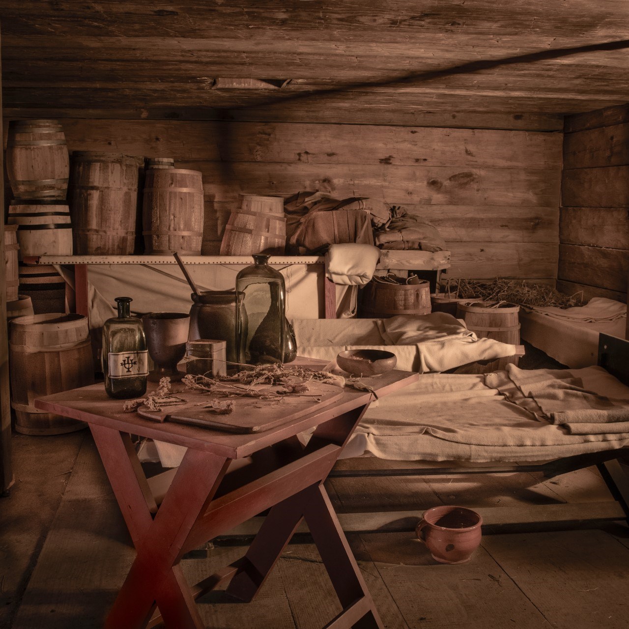 A dark room with cots and blankets, barrels stacked behind them.