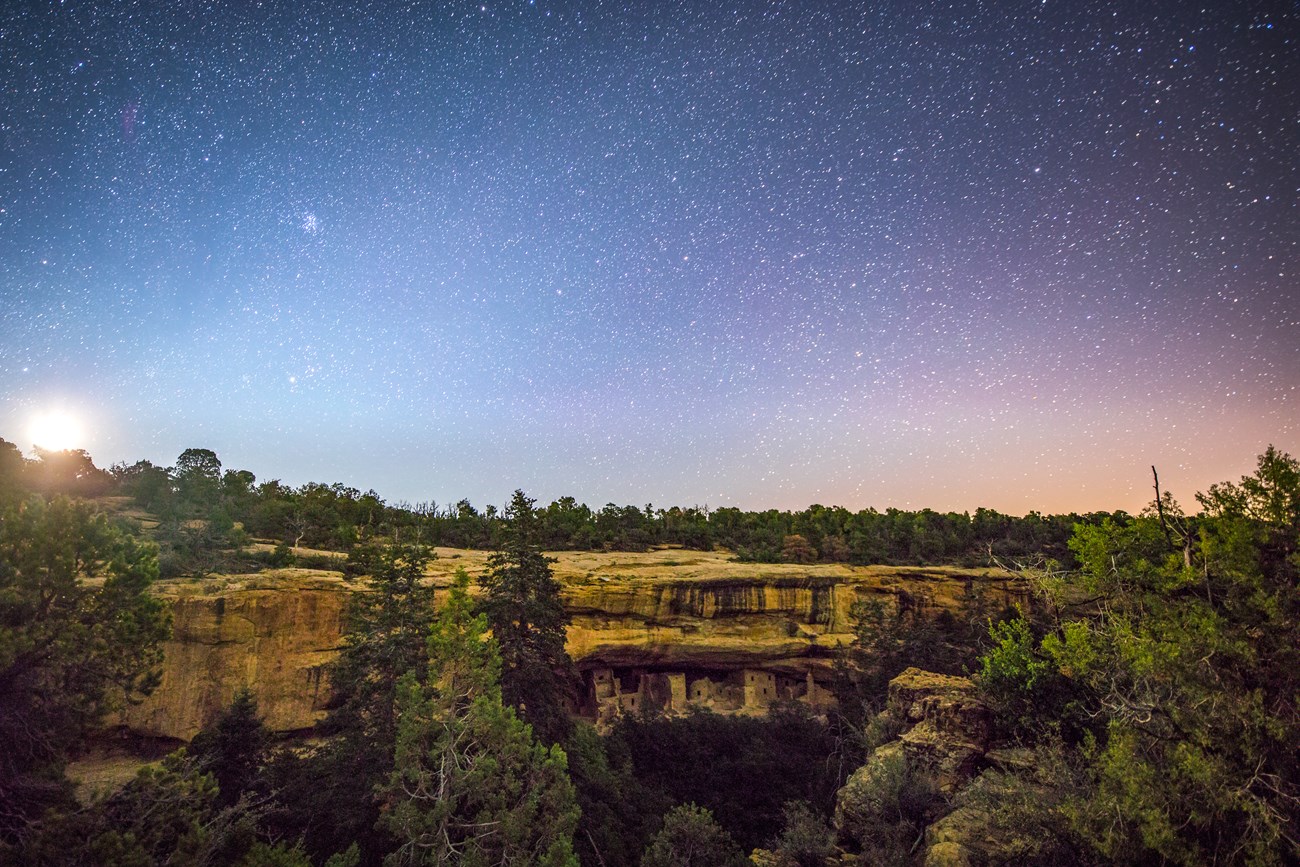 A full moon rising into a starry sky over a stone village built into a cliff alcove in a forested canyon.
