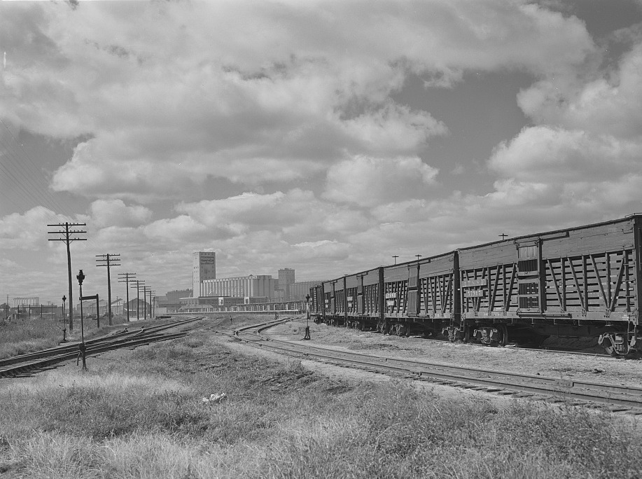 Railroad tracks and a line of slatted stock cars beneath a big sky, with grain elevators and a flour mill on the skyline
