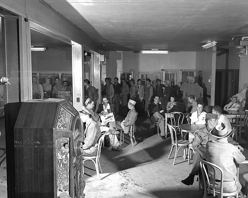 A crowded wartime cafeteria with people seated at small tables and a line of workers along the far wall, a jukebox in the foreground