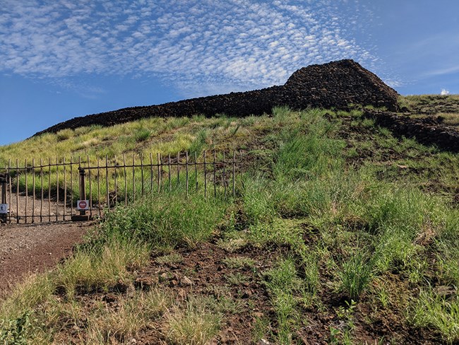 The stone platform of Puʻukoholā Heiau on a hill above strips of green grass, seen from the battlefield area behind an iron fence