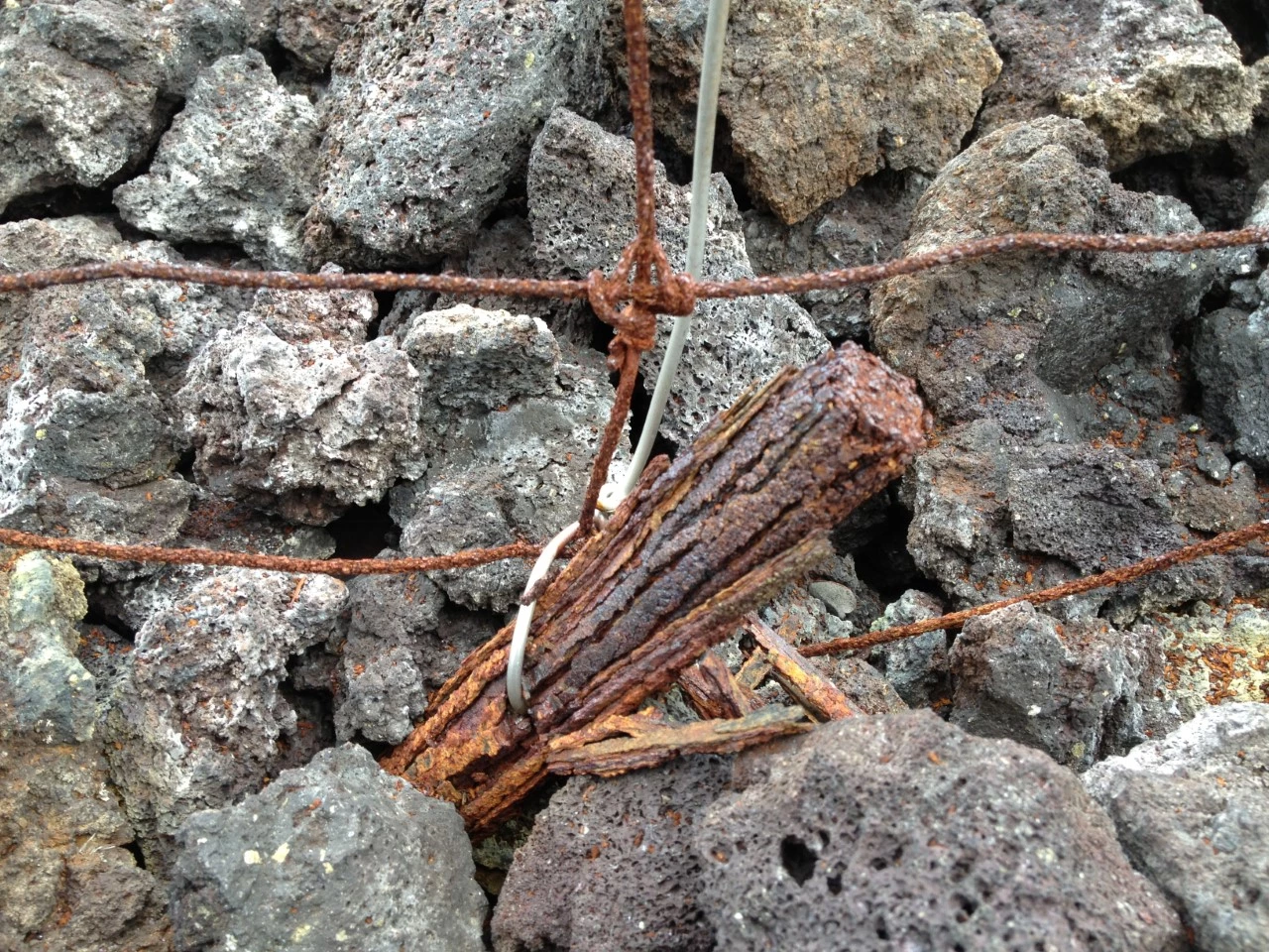 A corroded fence anchor wrapped in rusty wire, jammed among gray rocks