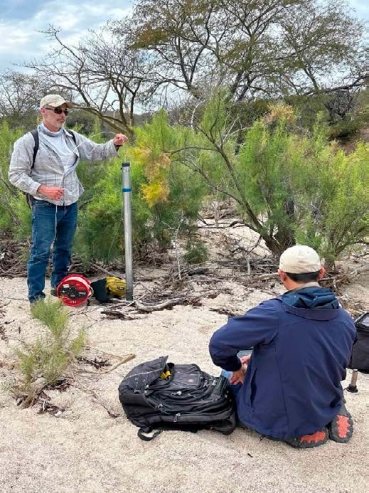 Two men beside a monitoring well in a sandy, shrubby streambed