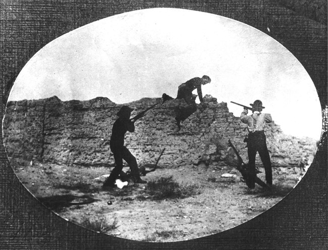 Men with rifles next to the remains of an adobe wall.