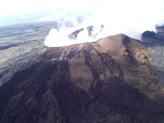 An aerial view of a fuming volcanic cone with a steam plume on the distant coast