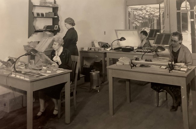 Three women at desks with lighted magnifiers while a fourth stands at a shelf