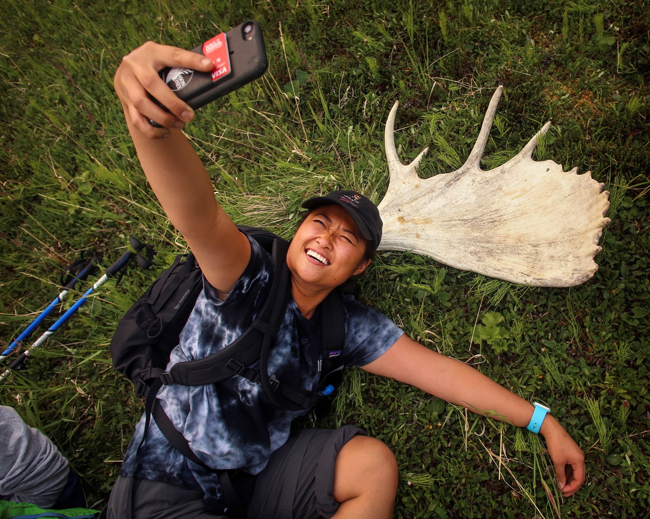 A hiker lying on the grass taking a selfie beside a shed moose antler.