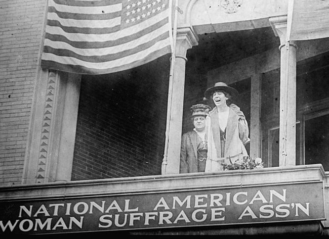 Jeannette Rankin speaking from the balcony of the National American Woman Suffrage Association’s headquarters beneath an American flag, with Carrie Chapman Catt behind her.