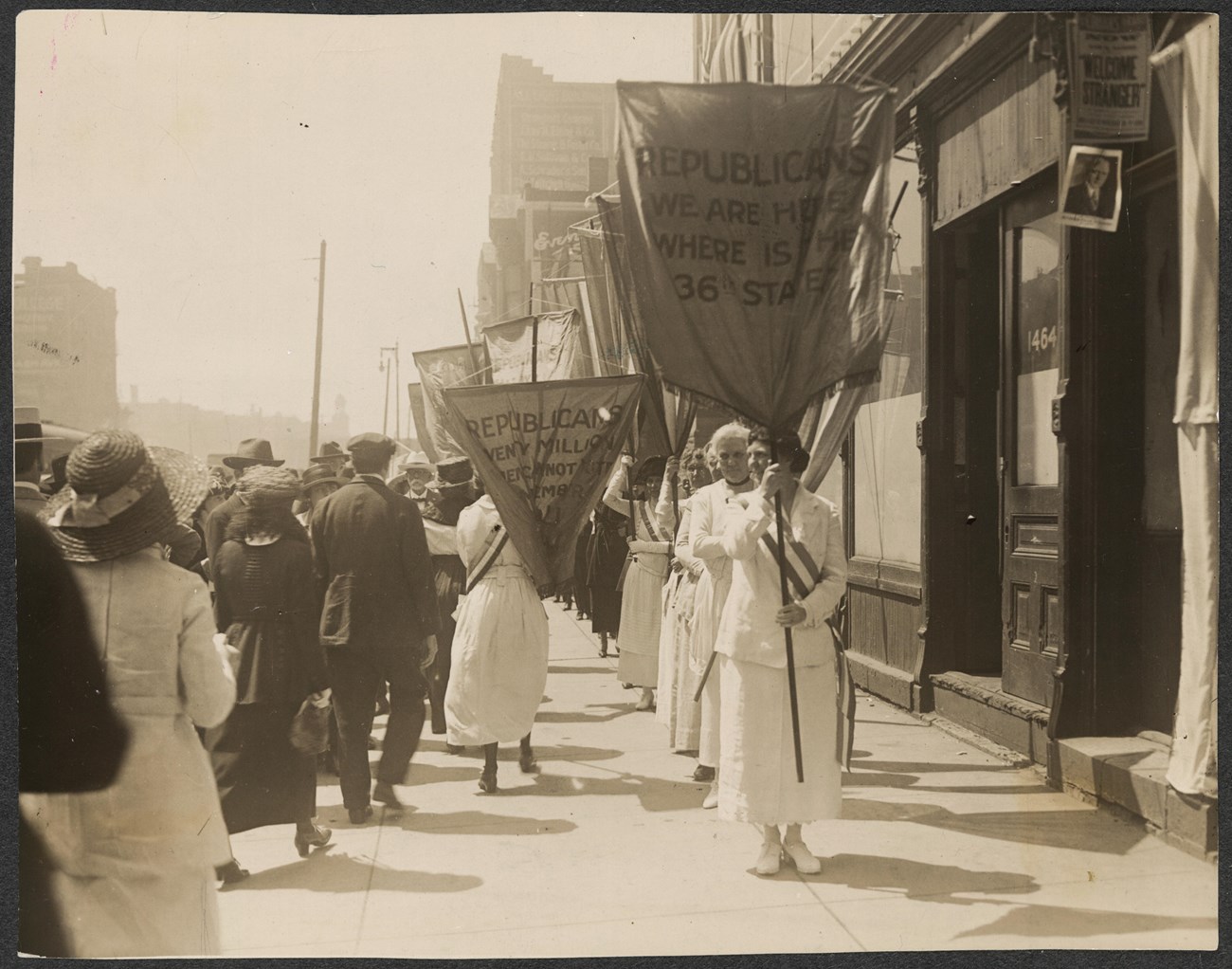 Suffragists picketing on a city sidewalk with banners, one reading "Republicans We Are Here Where is The 36th State?"