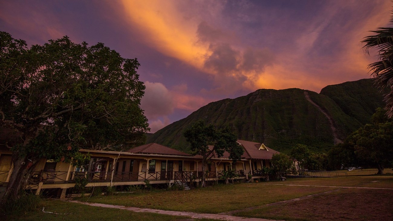 A long, low brown building with a continuous porch among tropical trees at the foot of a green mountain at sunset.