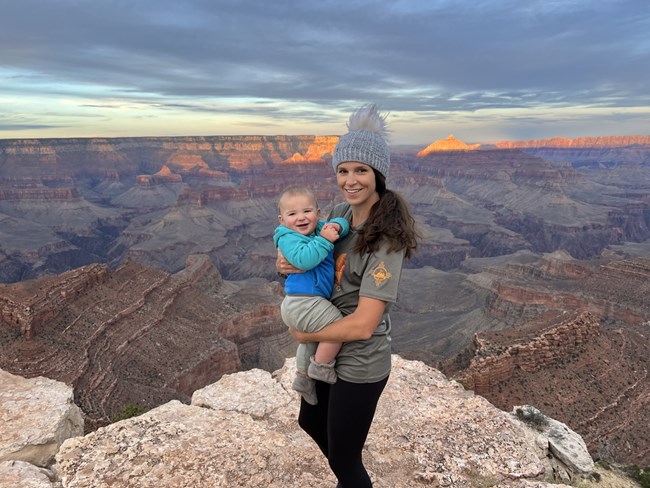 Jen Baker stands with a small child in her arms on a vista with a Grand Canyon sunset behind her.