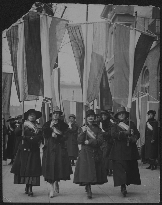 Four women in coats, hats and suffrage sashes carrying tricolor banners on a city sidewalk.