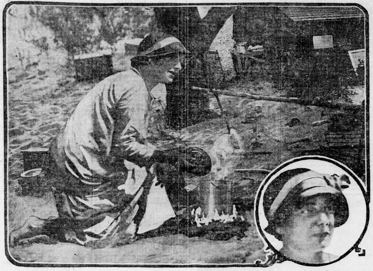 A woman in a long light-colored garment and a hat bends over a campfire, tending a pot, with sand, trees and wooden structures behind her.
