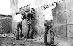 Three workers using tools to strip cement plaster from the church's exterior wall