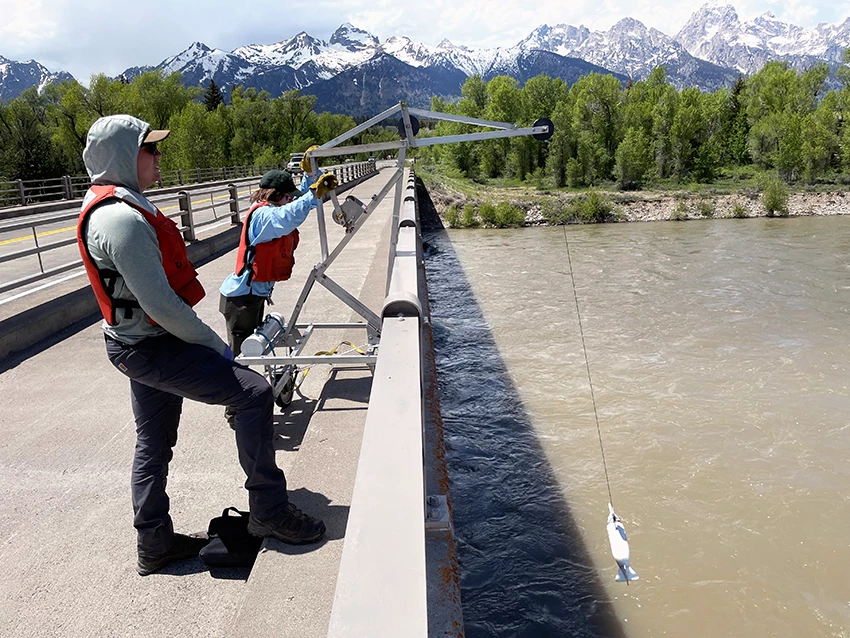 Two people on a bridge operate a crane and reel to lower a sampler into the river.