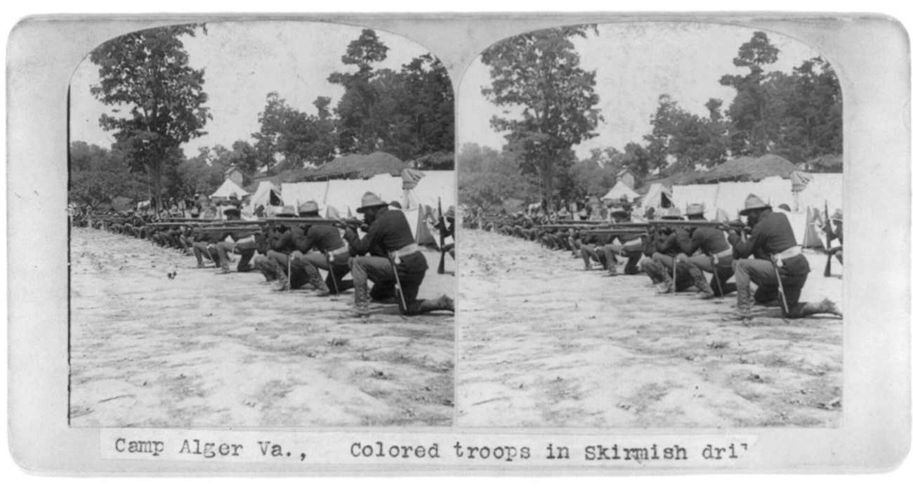 A stereograph of African American soldiers kneeling in a long line, rifles at their shoulders, with tents behind