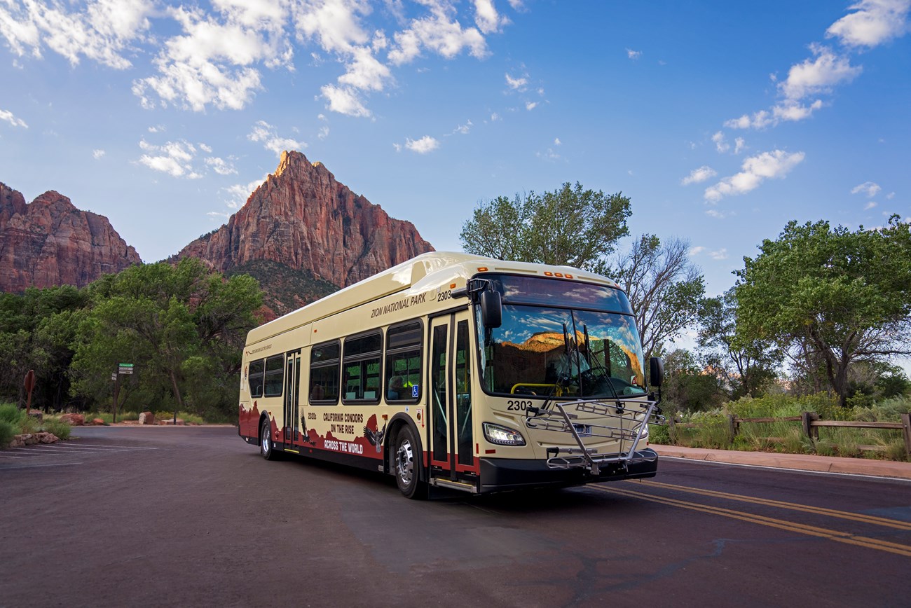 A beige shuttle bus on a road in front of a huge, triangular sandstone peak.