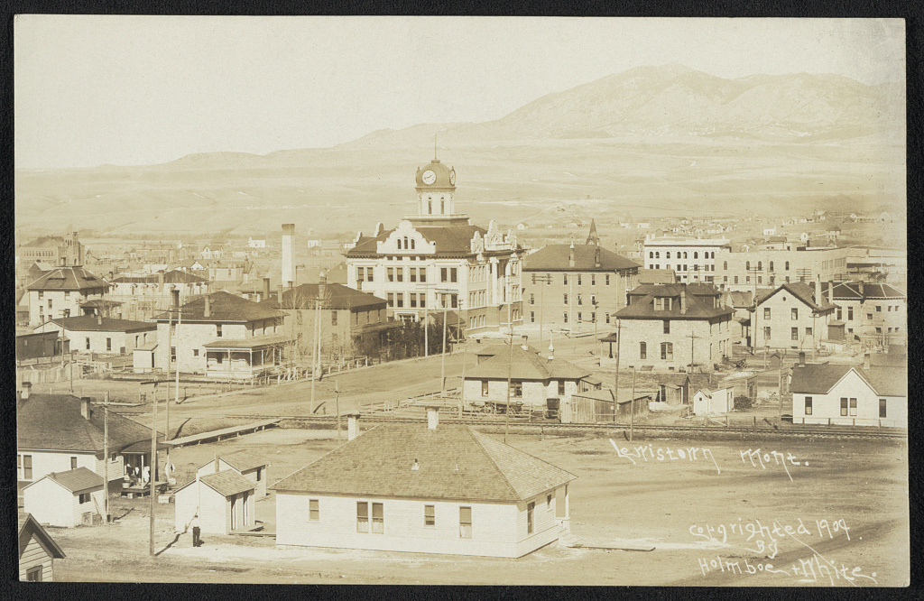 Old photograph of a small town on a plain: houses and a large courthouse with a clock tower, with mountains beyond