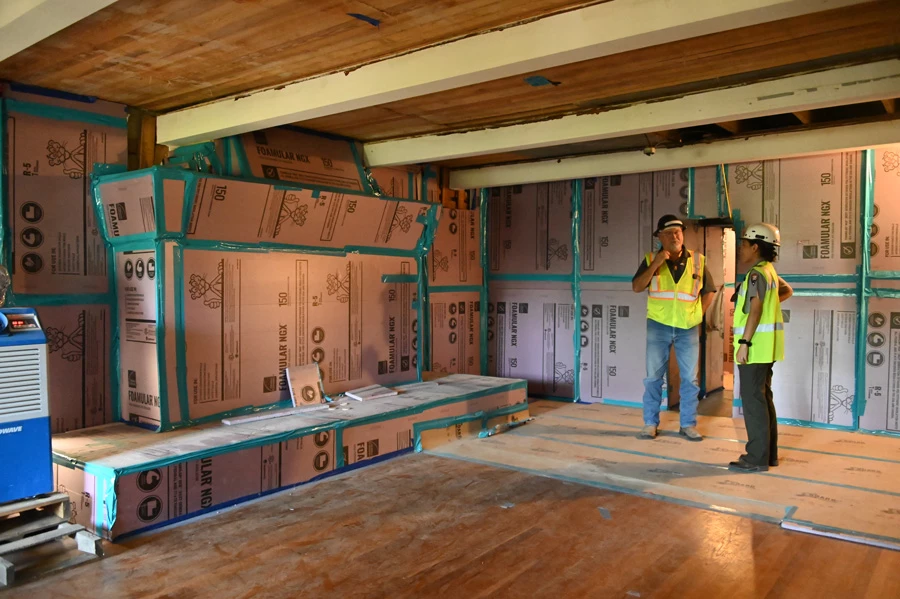 Two people wearing construction helmets stand inside a room with wood floor and ceiling, walls hidden behind protective covering.
