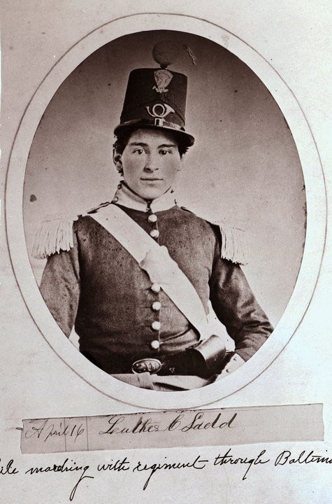 A black and white portrait of a young man wearing an intricate unifrom. Below the words read 'April 16, Luther C Ladd, Marching with regiment through Baltimore'.