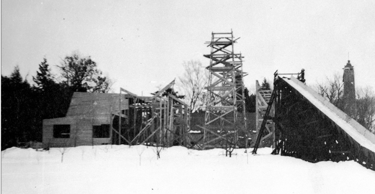 Bare orchard trees in snow beside a building and a toboggan chute under construction, 1904.