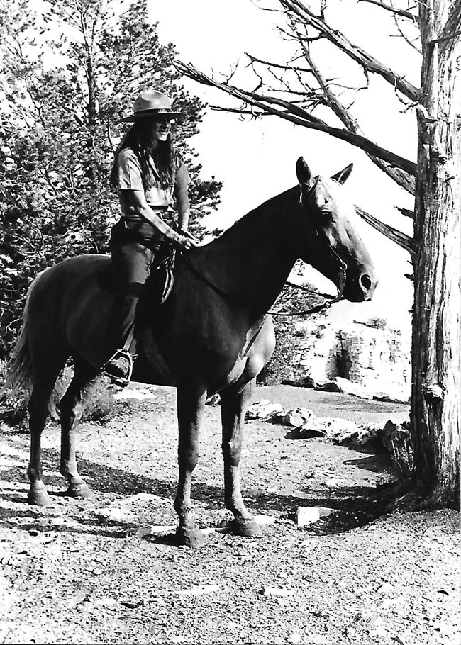 A ranger in uniform and flat hat riding a horse at the Grand Canyon.