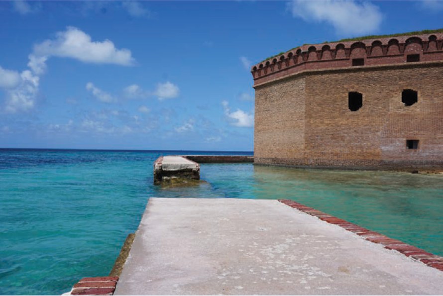 A broken concrete wall in turquoise water beside a brick fort