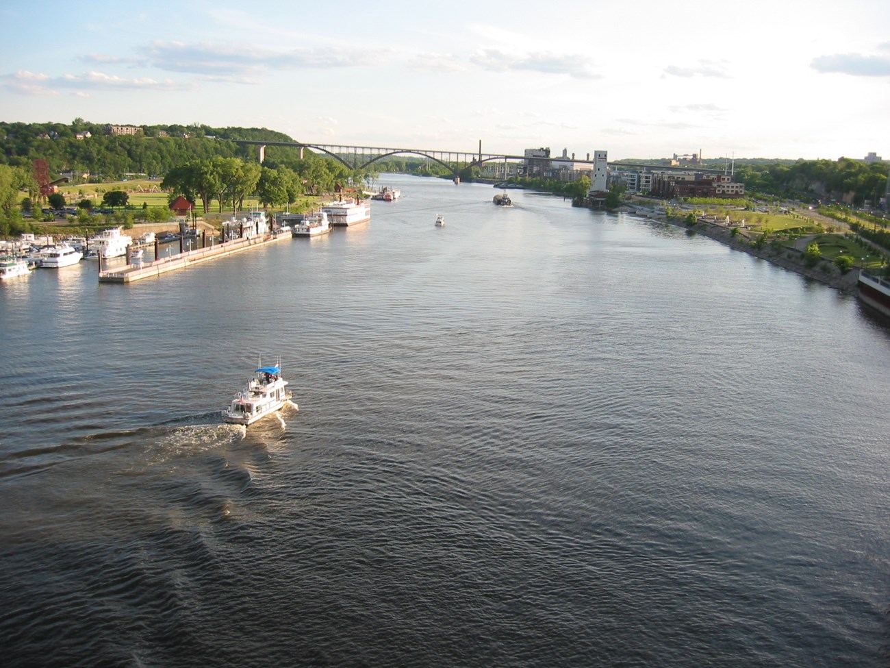 Boats on a river lined with grassy parks, buildings and a road bridge.