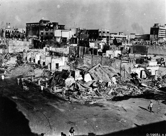 Black and white photo overlooking ruins of buildings. In the foreground, only a few walls are standing. In the distance, windows are blown out and there is evidence of fire.