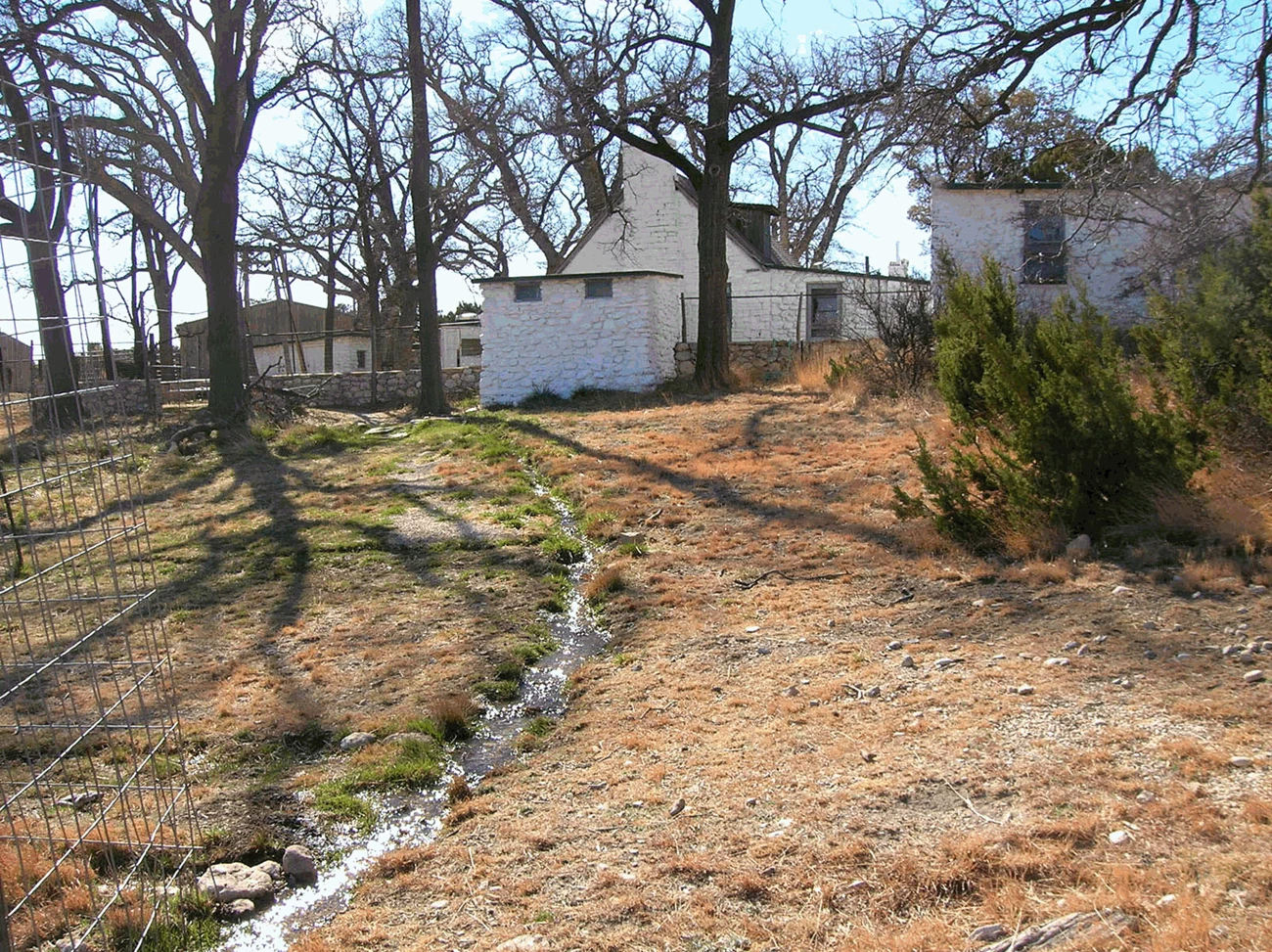 Water flows down a slope through an irrigation channel, looking toward the Ranch House cluster and leafless trees.