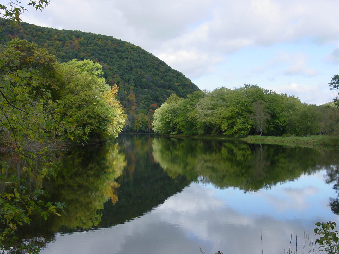 A calm river reflecting a wooded hill and early fall trees under a cloudy sky