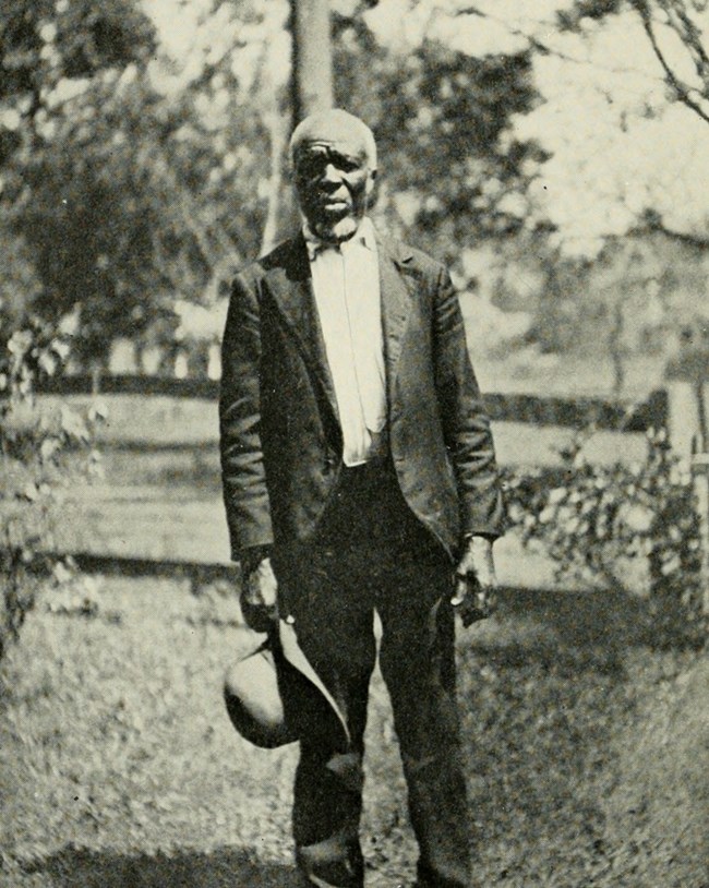 A Black man in a suit stands outdoors holding his hat.