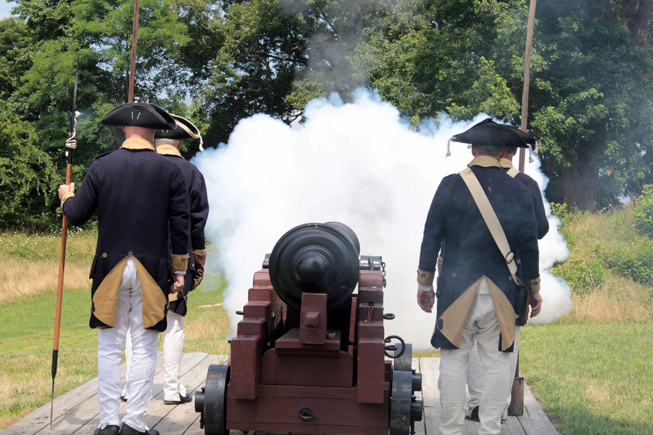 Reenactors in Continental uniforms fire a field cannon, a cloud of white smoke billowing from the muzzle